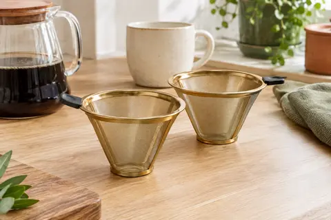 Deux filtres à café réutilisables à maille dorée posés près d’une cafetière en verre et d’une tasse