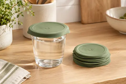 Couvercles extensibles verts en silicone, dont un posé sur un verre dans une cuisine lumineuse.
