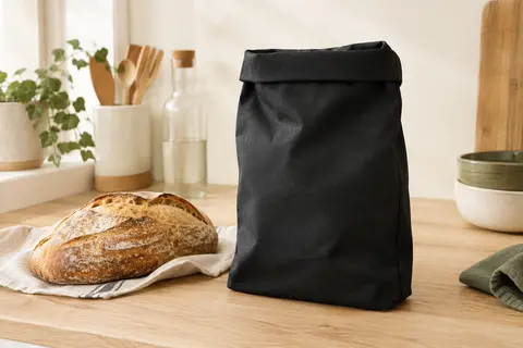 Sac à pain souple en polyester noir posé sur un plan de travail en bois clair, près d’un pain.