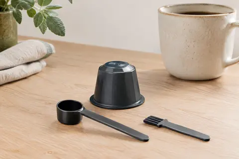 Capsule de café rechargeable en plastique PP avec sa cuillère et son pinceau sur un plan de travail.