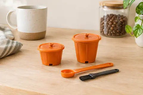 Deux capsules de café réutilisables orange accompagnées d’une cuillère et d’un pinceau sur un plan de cuisine clair.