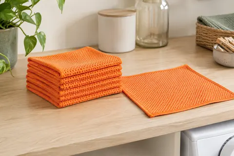 Lot de lavettes microfibres orange pliées, avec leur texture en nid d’abeille visible sur un plan en bois clair.